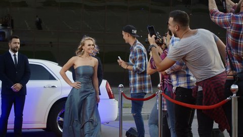 Cheerful blonde woman in gray dress getting out of limousine on red carpet while bodyguards stop the fan