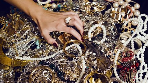 Female hand with a large ring of gold with precious stones touches a pile of gold and silver jewelry on a black background. Luxurious life. Incredible wealth. Hidden treasures.