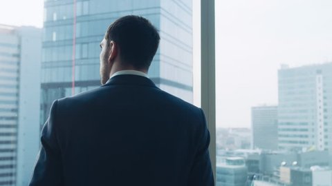 Medium Shot of Successful Businessman wearing a Suit Standing in His Office, Contemplating Next Big Business Deal, Looking out of the Window. Shot on RED EPIC-W 8K Helium Cinema Camera.