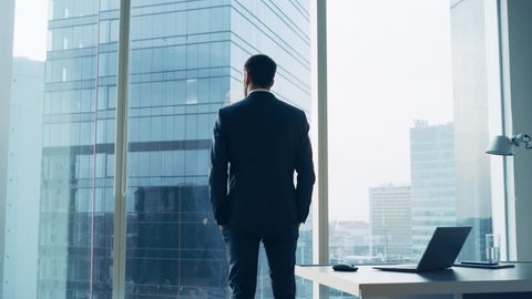 Back View of the Thoughtful Businessman wearing a Suit Standing in His Office, Hands in Pockets and Contemplating Next Big Business Deal, Looking out of the Window. 