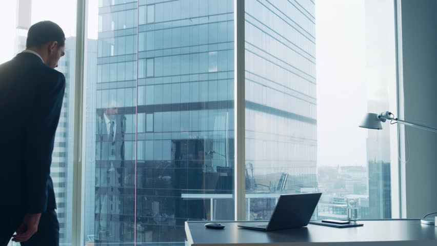 Following Shot of the Confident Businessman in a Suit Walking Through His Office and Leaning Against Window Frame, Looking out of the Window Thoughtfully.  Royalty-Free Stock Footage #1014231623