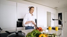 Happy pretty blonde woman in headphones cooking vegetables while listening music and dancing on kitchen at home