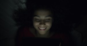 Profile of Beautiful Mixed Race Teen Curly Afro Girl on Touch screen Smart Pad in Dark Room with Mobile Night Light, Young Happy Woman Student Watching Social Media in the evening.