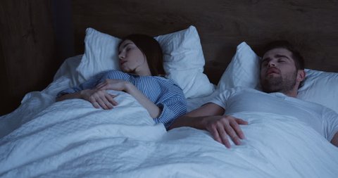 Caucasian couple sleeping in the bed at night. Woman waking up and closing ears with hands as man snoring, then trying to wake him up.