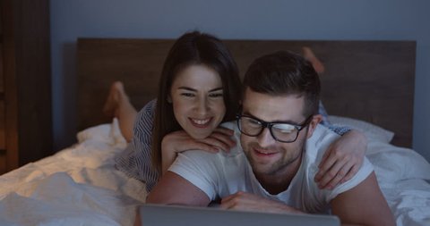 Close up of the young attractive couple lying on the bed in the evening and watching something funny on the laptop computer.