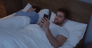 Young handsome Caucasian man lying in the bed near his young wife and cheating on her while chatting on the smartphone with another woman late in the night.