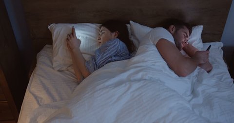 Top view on the married young Caucasian couple lying in the bed back to back at night and taping or scrolling on smartphones. Indoor. Stock-video