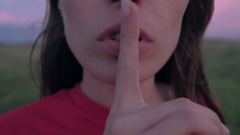 Close-up shoot of attractive woman gesturing for silence or telling secret. She raises finger to her lips.