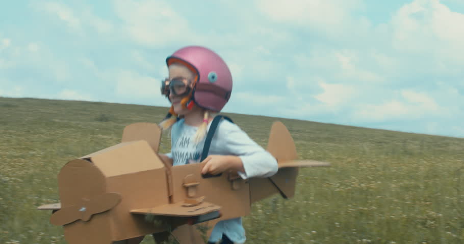 TRACKING Cute little dreamer kid girl wearing pink helmet and aviator glasses flying in a cardboard airplane through the field, pretending to be a pilot. 4K UHD 60 FPS SLO MO  Royalty-Free Stock Footage #1014267215