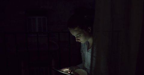 Young Woman swiping Smart Phone touch screen Device for social media behind Curtains at Night. Student Girl in Evening Light. A Bohemian Millenial