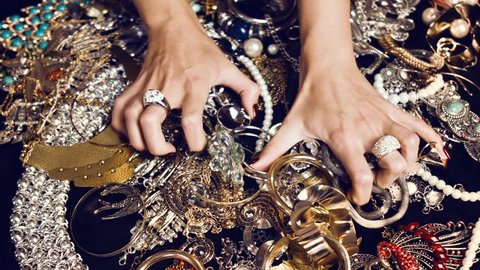 Female hands with a large ring of gold with precious stones touch a bunch of gold and silver jewelry on a black background. Luxurious life. Incredible wealth. Hidden treasures.