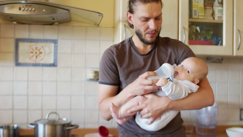 Son and father. A mature man feeds his young son from a bottle, standing in the kitchen near the stove. He smiles tenderly at the child Royalty-Free Stock Footage #1014270104