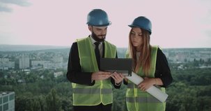 Two young engineers man and female have a discussion on the top of construction site , using a tablet analyzing the plan of building.