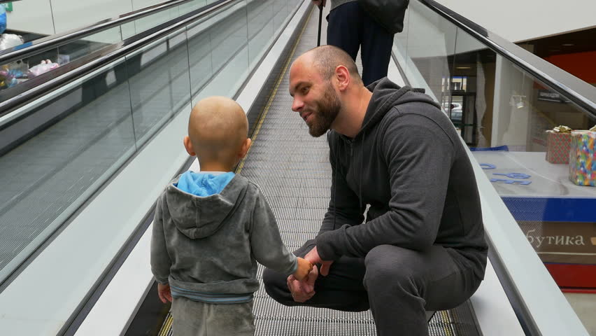 Back view of dad and his little son riding escalator in a shopping center