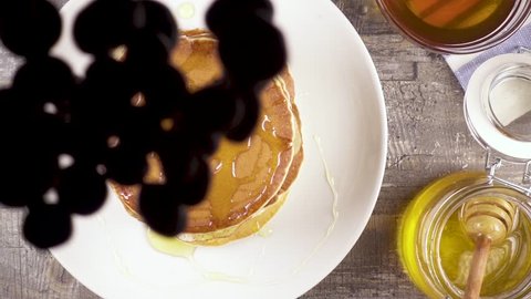 Slow motion blueberries falls on a stack of pancakes with honey on a white plate top view
