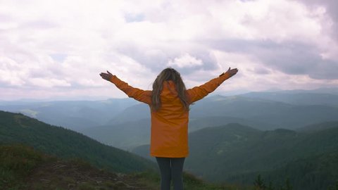 Unidentified young woman in yellow coat raising hands and looking at the beautiful mountains, back view