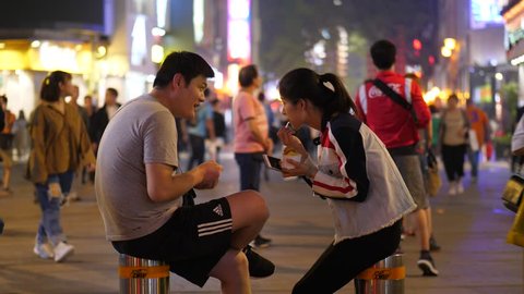 GUANGZHOU, CHINA - MARCH 16, 2018: Young Unidentified Asian Couple sit with ice cream in middle of pedestrian street, lively area at night time. People pass by, bright Beijing Lu shopping area 