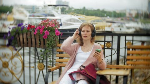 A girl using a smartphone on a bench in flower on the beach, listening to music in headphones, dancing a yacht and sailing in the harbor...4k