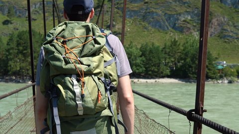 A tourist with a big green rucksack is walking along a suspension bridge that hangs over the river. Back view. Video de stock