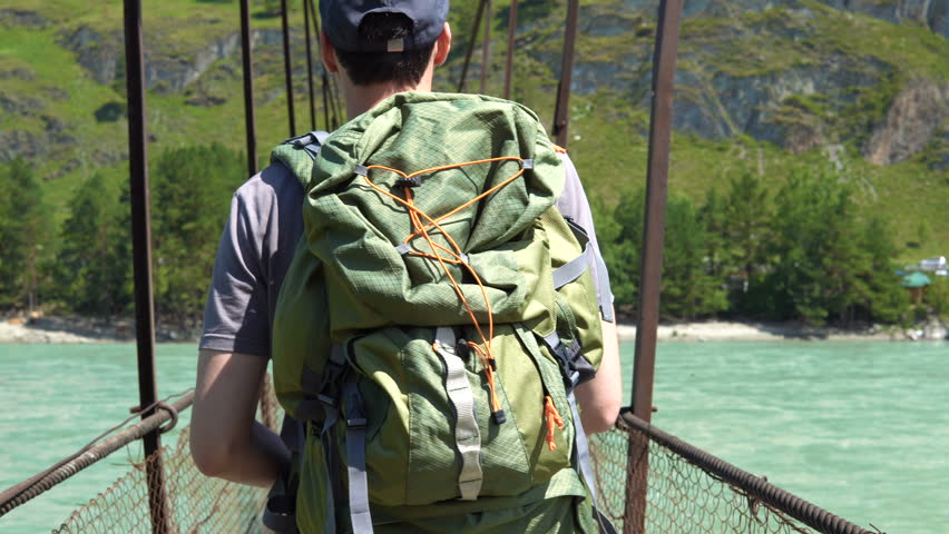 A tourist with a big green rucksack is walking along a suspension bridge that hangs over the river. Back view.