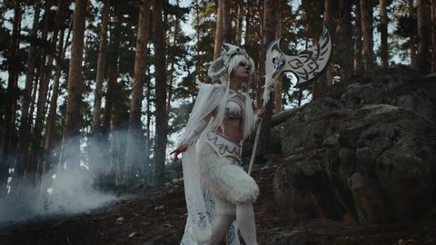 Model dressed like mythical faun is strolling in forest near spruces. She is carrying hatchet in hands, tilt up view