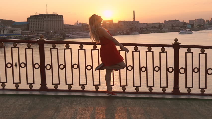 young cheerful girl dances over the bridge and looks at the sunset. hair bounce in slow motion. red chiffon dress on a woman. Sunlight is reflected in the water