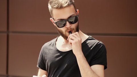A young handsome white bearded man with smartphone is vaping an electronic cigarette in the street in the spring. Close up.