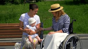 Mom, child and girl in a wheelchair, sit side by side, smile and talk. 
Two women and a child walk in the park