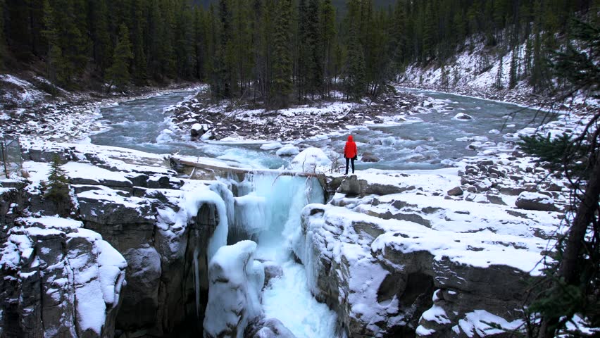 Man in orange standing at edge of waterfall in nature. Sunwapta Falls, Alberta, Canada