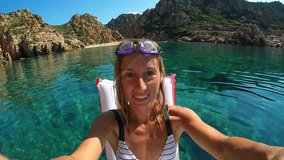 Young woman taking selfie portrait with an action camera while floating on water with an inflatable mattress. Shot in Sardinia, Italy in the Mediterranean Sea. People vacations fun summer concept