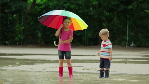 Little boy and girl play in rainy summer park. Children with waterproof boots jump in puddle and mud in the rain. Kids walk in autumn shower. Outdoor fun by any weather.
