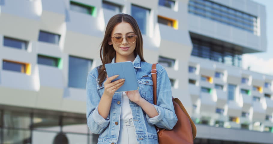 Caucasian young good looking woman with long hair typing while texting on the tablet computer at the big urban building background. Outside.