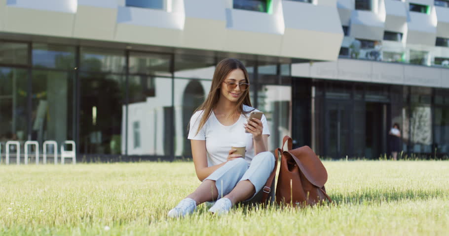Caucasian young pretty woman in the white T-shirt using her smartphone and drinking coffee on the modern building background. Outdoors.