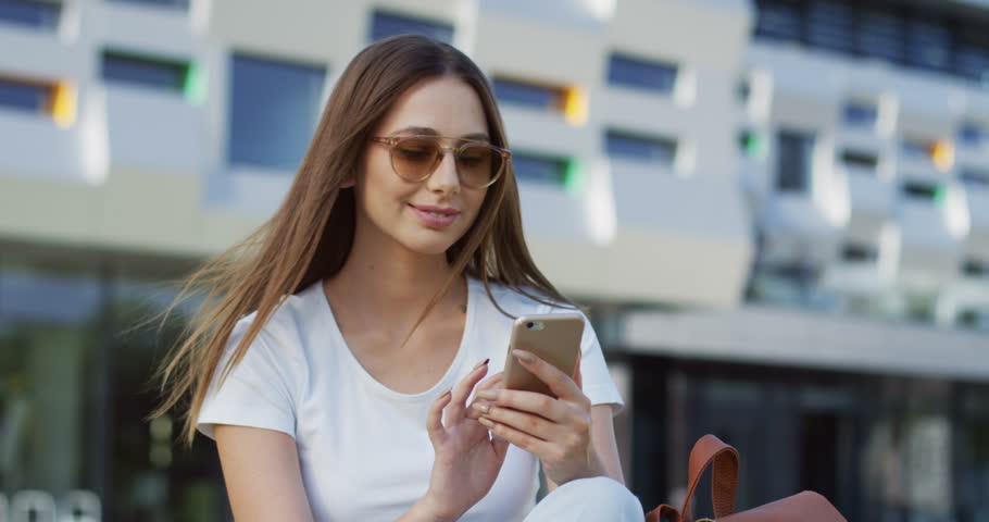Close up of the young charming woman in the white T-shirt scrolling and taping on the smartphone on the modern building background. Outside.