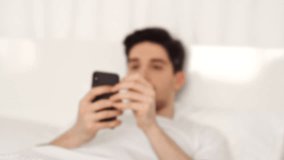 Happy surprised brunette smiing man wearing casual clothes lying in bed and using smartphone
