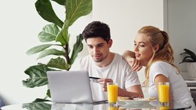 Smiling loving couple having breakfast while sitting at the table in a kitchen at home and looking at laptop computer and using credit card