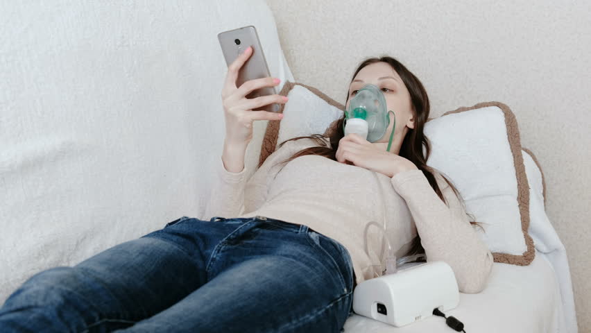 Use nebulizer and inhaler for the treatment. Young woman inhaling through inhaler mask lying on the couch and chatting in mobile phone. Side view.