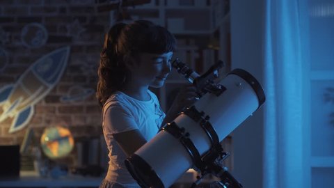 Cute girl watching the stars with a professional telescope at night in her room, imagination and childhood concept