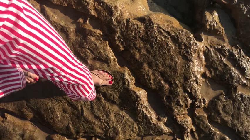 Female legs red nail polish toes and red white striped pants walking stones shore reaching sea water swash