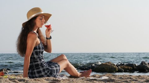 Young beautiful woman in hat drinking wine near sea on the beach. Girlfriend waiting for her man on picnic. Travel, relax concept. Slow motion