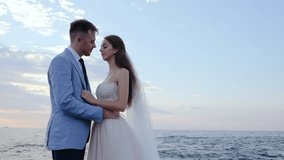 Beautiful young wedding couple standing on sea shore with rocks. Newlyweds spend time together: embrace, kiss and care for each other. Love concept. Bride smiling to groom.
