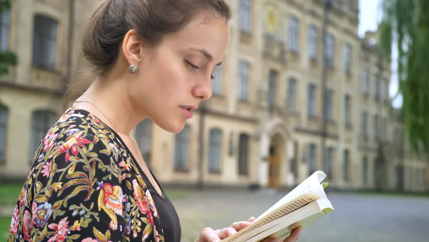 Profile of young beautiful woman reading book and sitting on street near college building, concentrated and determined