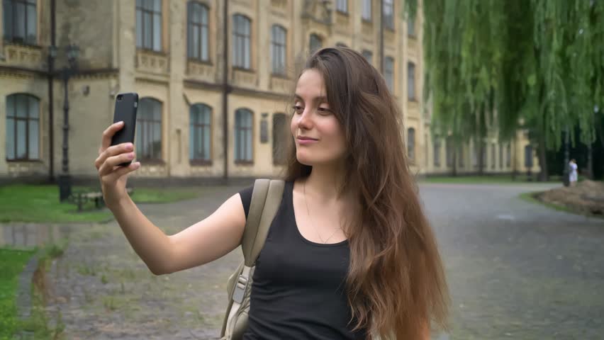 Young charming woman with long hair having video chat through phone, smiling, standing near college campus outdoors