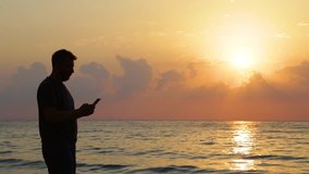 Black silhouette of adult tall man taking pictures of beautiful scenic seascape and selfie during sunrise on early morning. Real time full hd video footage.