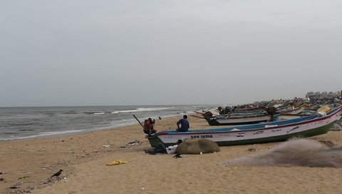 Chennai,India,July 15 2018 handheld unedited footage of fishing boats parked in line near seashore,marina beach,with unidentified people having leisure time in sea breeze,beautiful waves.Low light.