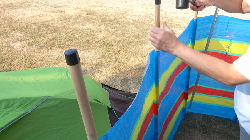 POV close shot of a man’s hands securing wooden windbreak poles next to a small tent on a campsite, by hammering the tops with a rubber mallet.