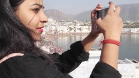 Handheld woman takes selfie photo video with her mobile cellphone device from a panoramic vantage view point of holy Pushkar lake, a sacred city for Hindu pilgrims mela festival bathing ghats temples