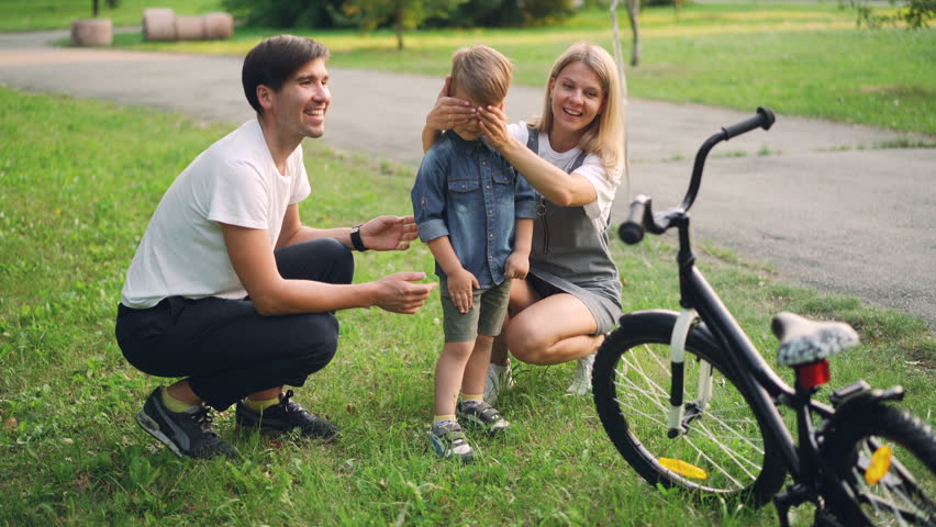 Loving parents are making surprise for little son closing his eyes and giving him new bicycle as present, happy boy is looking at bike and talking to mother and father.