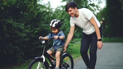 Slow motion of excited boy riding bicycle and laughing while his careful father is helping him holding bike and teaching child to ride. Family, sports and childhood concept.