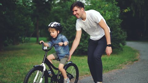 Slow motion of laughing child cycling in park with careful father who is teaching him to ride bicycle. Happy young family, fatherhood and childhood, active lifestyle concept.